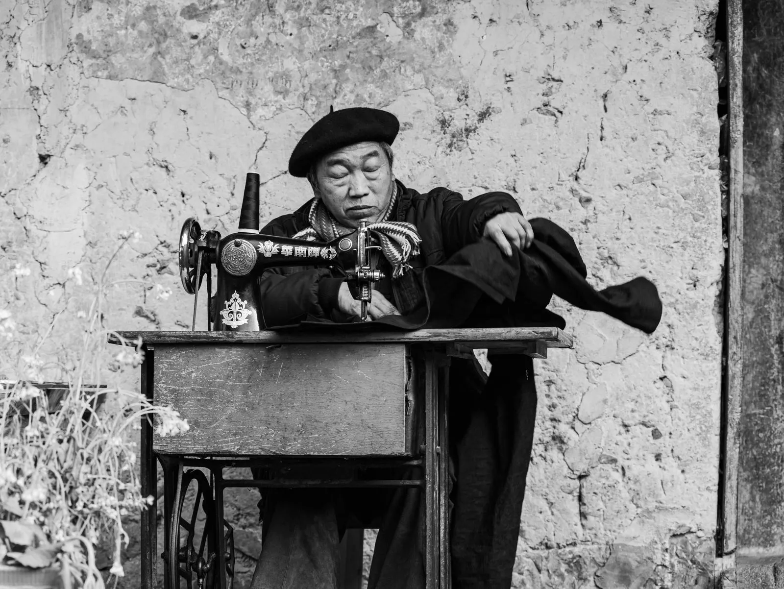 Black and white photograph of a tailor working with a vintage sewing machine outdoors.